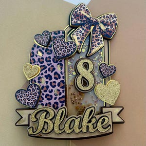 Puede incluir: Un adorno para pastel personalizado con un lazo con estampado de leopardo, corazones y el nombre "Blake" en purpurina dorada. También aparece el número "8". El fondo tiene una combinación de colores rosa claro y dorado.
