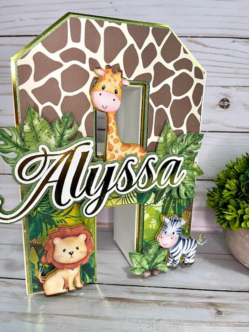 K&ouml;nnte beinhalten: Ein Dschungel-Tortenaufsatz mit einer braunen und wei&szlig;en Giraffe, einem L&ouml;wen und einem Zebra. Der Name "Alyssa" ist in goldenen Buchstaben geschrieben. Der Tortenaufsatz ist aus Pappe und hat einen Giraffenmuster-Hintergrund.