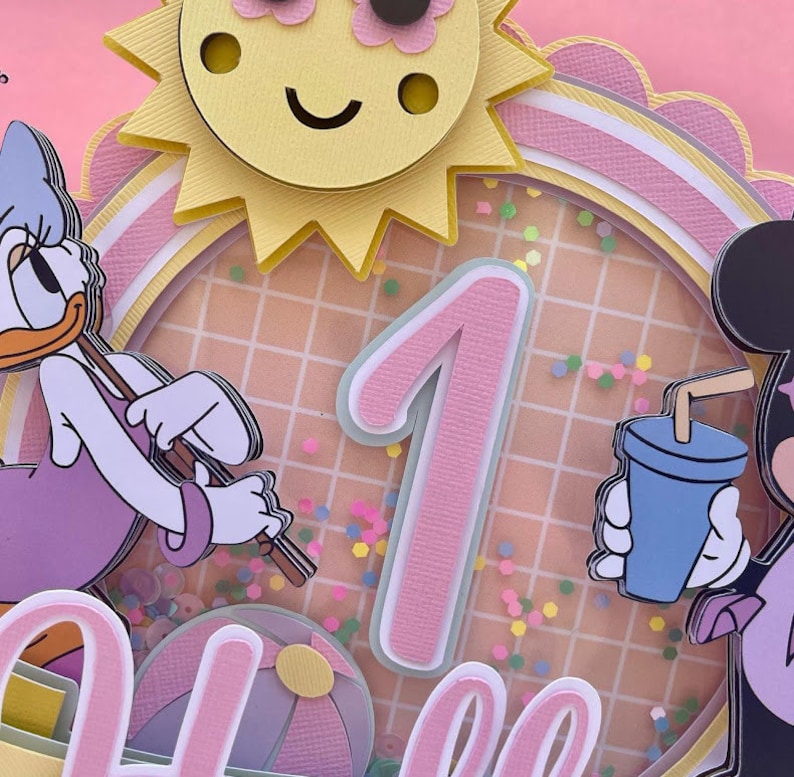 K&ouml;nnte beinhalten: Eine rosa und wei&szlig;e mehrst&ouml;ckige Torte mit einer Sonne, einer Eins und einer Mickey-Maus-Figur. Die Torte ist mit bunten Streuseln und einem rosa-wei&szlig;en Karomuster verziert.