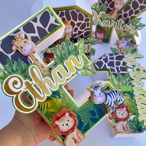 K&ouml;nnte beinhalten: Ein Set aus drei Dschungel-Kuchen-Toppern, die jeweils einen Buchstaben des Namens "Ethan" in einer wei&szlig;en Pappbox mit einem gr&uuml;nen, braunen und goldenen Tierdruckdesign zeigen. Die Buchstaben sind mit gr&uuml;nen Bl&auml;ttern, einer Giraffe, einem Zebra und einem L&ouml;wen verziert.