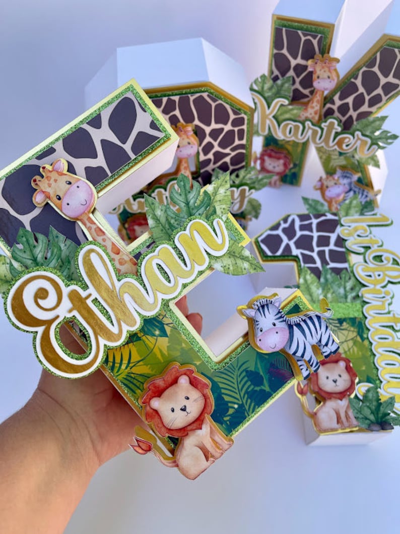 K&ouml;nnte beinhalten: Ein Set aus drei Dschungel-Kuchen-Toppern, die jeweils einen Buchstaben des Namens "Ethan" in einer wei&szlig;en Pappbox mit einem gr&uuml;nen, braunen und goldenen Tierdruckdesign zeigen. Die Buchstaben sind mit gr&uuml;nen Bl&auml;ttern, einer Giraffe, einem Zebra und einem L&ouml;wen verziert.