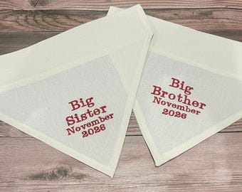 May include: Two white, triangular bandanas with red embroidered text. One reads "Big Sister November 2026" and the other "Big Brother November 2026". The bandanas are laid on a wooden surface.