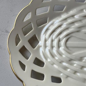May include: Close-up of a cream-colored, open-weave ceramic basket with a gold rim. The basket has a scalloped edge and a lattice design, with a textured interior. The basket is likely used for serving or display.