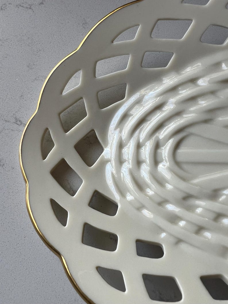 May include: Close-up of a cream-colored, open-weave ceramic basket with a gold rim. The basket has a scalloped edge and a lattice design, with a textured interior. The basket is likely used for serving or display.