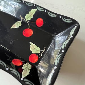 May include: Black rectangular serving tray with a glossy finish. The tray is decorated with red cherries and green leaves. The edges feature a decorative pattern in white and green. A decorative piece for serving food.