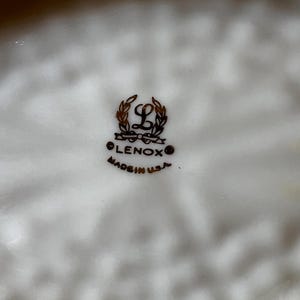 May include: Close-up of a white Lenox plate with a decorative rim. The Lenox logo, featuring a stylized "L" within a wreath, is centered. The text "LENOX" and "MADE IN U.S.A." are below the logo.