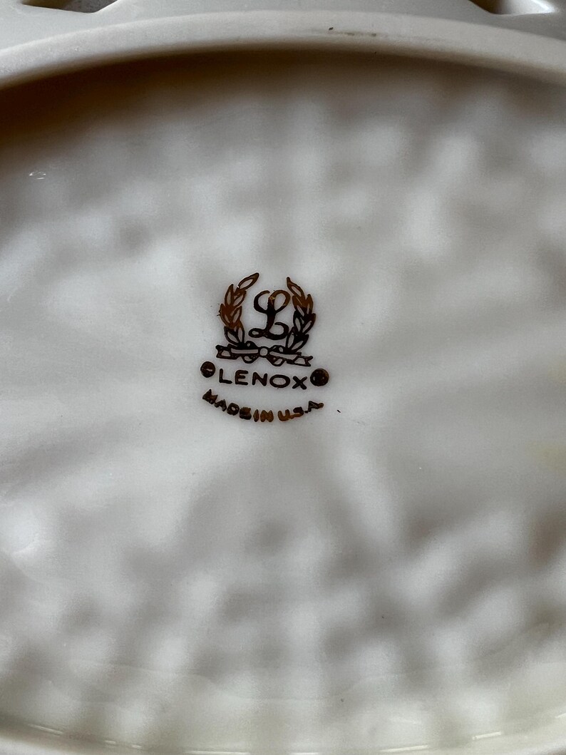 May include: Close-up of a white Lenox plate with a decorative rim. The Lenox logo, featuring a stylized "L" within a wreath, is centered. The text "LENOX" and "MADE IN U.S.A." are below the logo.