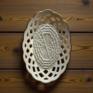 May include: Oval-shaped, white ceramic basket with a woven center and openwork sides. The basket has a gold-colored trim around the edge. The basket is designed to hold small items and is placed on a wooden surface.