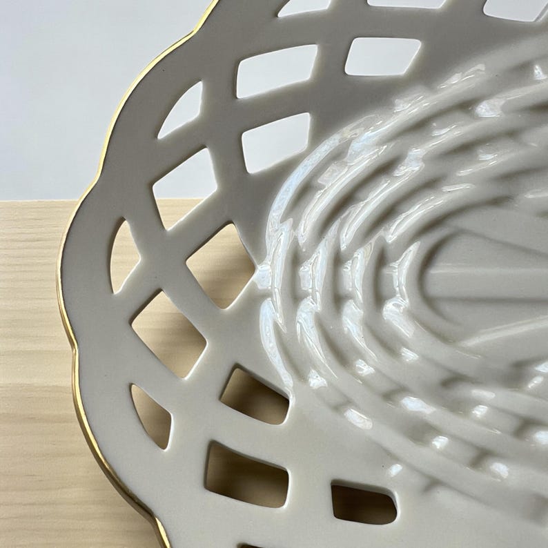 May include: A white ceramic serving dish with a lattice design and a gold rim. The dish has a detailed, openwork pattern and a smooth, glossy finish. The design is symmetrical and elegant, suitable for serving food or as a decorative piece.