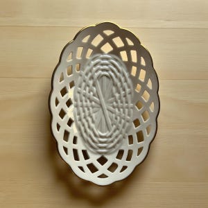 May include: An oval, white ceramic basket with a lattice design and a gold rim. The interior features a woven pattern. This decorative piece is ideal for displaying small items or as a decorative accent.
