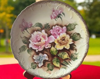May include: A decorative porcelain plate featuring a hand-painted floral design. The plate showcases pink, yellow, and blue roses with green leaves against a cream-colored background. The rim has a gold-colored trim and a scalloped edge. The plate is displayed on a wooden stand.