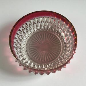 May include: A clear glass bowl with a ruby red rim and a gold-colored band. The bowl has a diamond-cut pattern on the sides and a starburst design on the bottom. The bowl is likely vintage and suitable for serving.