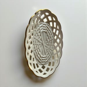 May include: An oval, white ceramic basket with a lattice design. The basket has a gold-colored trim around the edge. The interior of the basket has a woven pattern. The basket is empty and sits on a white surface.
