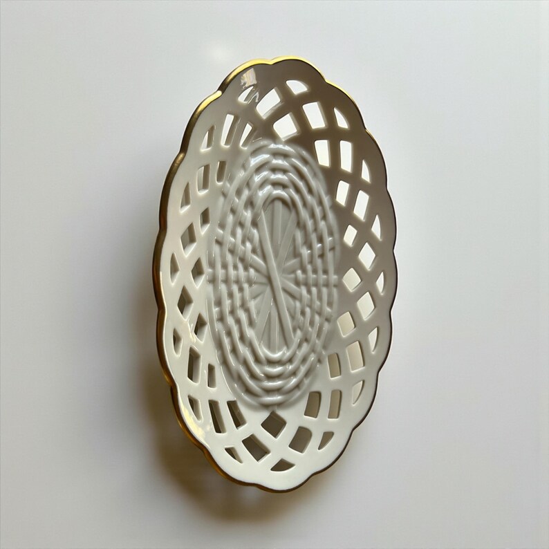 May include: An oval, white ceramic basket with a lattice design. The basket has a gold-colored trim around the edge. The interior of the basket has a woven pattern. The basket is empty and sits on a white surface.