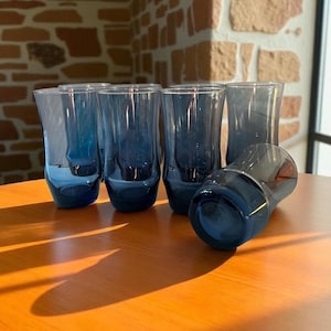 May include: Six blue-tinted drinking glasses are displayed on a wooden surface. The glasses have a curved design. The glasses are empty and appear to be made of glass.