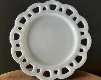 May include: A white, round decorative plate with a scalloped edge featuring openwork cutouts. The plate has a smooth, glossy surface and a slightly raised center. The design is symmetrical and the plate is likely made of ceramic or glass.
