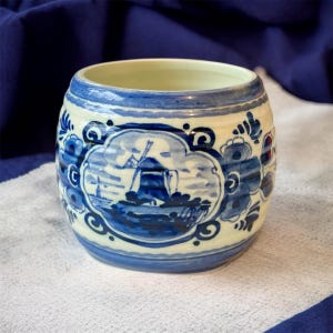 May include: A ceramic, barrel-shaped container with a blue and white windmill design. The container has a cream-coloured base with blue accents and a blue rim. The windmill scene is framed by a cloud-like border.