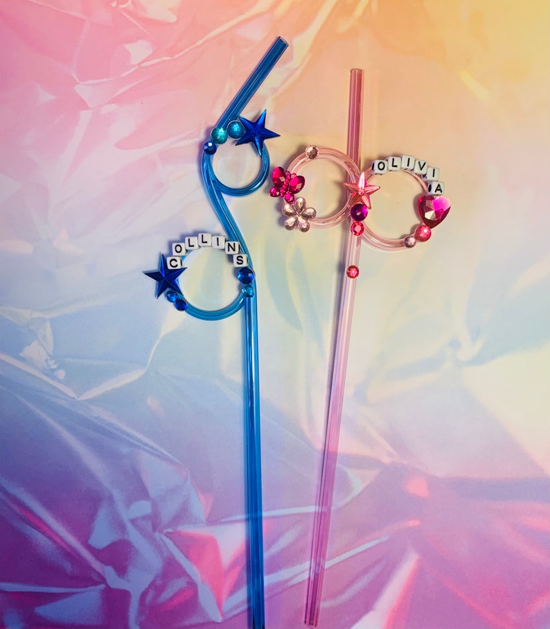 Puede incluir: Dos pajitas de pl&aacute;stico de colores con adornos decorativos. Una pajita es azul con una estrella y la palabra "COLLINS" escrita en letras blancas. La otra pajita es rosa con un coraz&oacute;n y la palabra "OLIVIA" escrita en letras blancas.