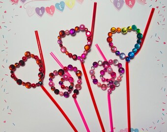 Puede incluir: Cinco pajitas rojas decoradas con cuentas de colores en forma de corazón. Las pajitas están dispuestas en forma de abanico sobre un fondo blanco con un patrón de confeti de corazones rojos, azules y blancos.