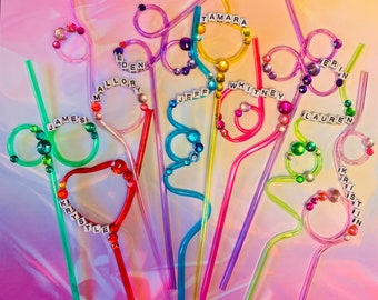 Puede incluir: Una colección de pajitas flexibles de colores con nombres escritos en letras blancas. Las pajitas están decoradas con joyas de colores.