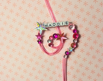 Puede incluir: Una varita mágica rosa personalizada con el nombre "MADDIE" escrito con cuentas de letras blancas. La varita está adornada con estrellas rosas e iridiscentes, y varias gemas rosas y transparentes. El fondo es un patrón rosa claro.