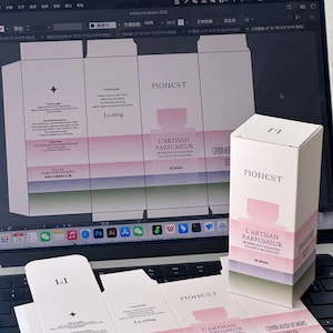 Può includere: Una scatola di profumo bianca e rosa con il testo "MONEST" e "L'ARTISAN PARFUMEUR". La scatola è su una tastiera di un laptop con un layout di design visibile sullo schermo e altri componenti della scatola.