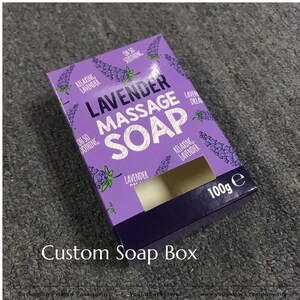 May include: A rectangular purple cardboard soap box featuring a lavender floral pattern and the printed text "LAVENDER MASSAGE SOAP", "RELAXING LAVENDER", "OH SO SOOTHING", and "LAVENDER DREAM". The box includes a rectangular cutout and displays 100g.