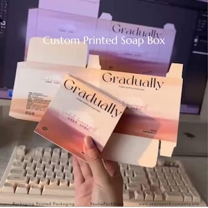 Puede incluir: Cajas de jabón impresas a medida con la palabra "Gradually" en una fuente moderna. Las cajas tienen un diseño degradado con tonos rosas y naranjas. También es visible el texto "Custom Printed Soap Box". El embalaje es para un producto de belleza.