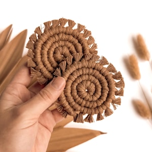 May include: Two brown macrame coasters with fringe details. The coasters are made with a woven rope and have a circular shape.