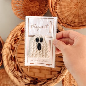 May include: A macrame ghost magnet featuring off-white twisted cotton cord and three black oval beads for eyes and a mouth. The ghost is attached to a rectangular display card with a refrigerator graphic and the text Magnet and @merakithestudios.
