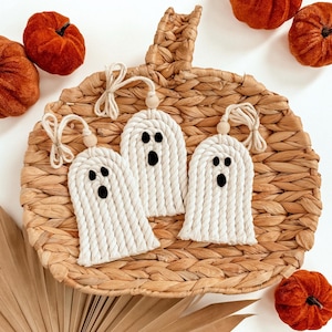 May include: Three handmade macrame ghost ornaments with black eyes and mouths, suspended from wooden beads and twine. They sit on a woven pumpkin-shaped tray, surrounded by orange velvet pumpkins, ideal for Halloween decorations.