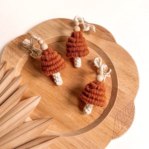 Op de afbeelding: Drie gehaakte paddenstoelversieringen in bruin en wit op een houten dienblad. De paddenstoelen hebben een textuur, geweven uiterlijk en zijn versierd met houten kralen.