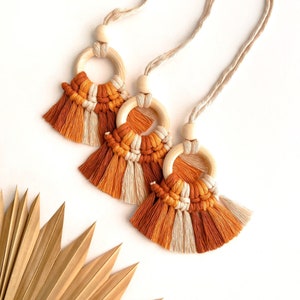 May include: Three macrame tassel keychains with wooden rings. The tassels are made with a combination of beige and orange yarn.