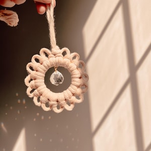 May include: A beige macrame ornament with a circular design and a dangling crystal. The ornament is suspended by a matching beige cord. The background is a neutral color with shadows.