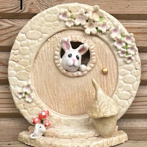 Puede incluir: Una caprichosa puerta de hadas con un diseño redondo de color crema. Un conejo blanco se asoma por la ventana de la puerta. La puerta está adornada con flores y una pequeña bellota. Un pequeño ratón blanco está sentado cerca de setas rojas y blancas.