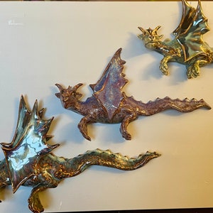 May include: Three ceramic dragons in flight, each with a different colour scheme. The dragons are brown, green, and purple with iridescent accents. They are all facing the right side of the image.