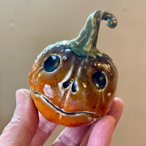 Puede incluir: Figura de calabaza de cerámica con una cara sonriente, ojos oscuros y un tallo curvo. La calabaza es principalmente naranja con acentos marrones y verdes, y tiene un acabado brillante. La figura se sostiene en una mano.