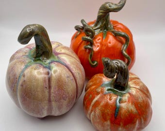May include: Three ceramic pumpkins with glossy finishes. One is a pale, mottled lavender and cream color, while the other two are vibrant orange with dark green, twisted stems and curling vine-like tendrils extending from the top of each gourd.