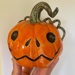 Può includere: Una zucca in ceramica dipinta a mano con uno stelo verde e tratti del viso neri. La zucca ha una glassa arancione lucida.