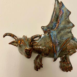 Op de afbeelding: Een keramisch drakenbeeld met groene en bruine glazuur. De draak heeft een brede vleugelspanwijdte en bevindt zich in een vliegende pose. De ogen van de draak zijn oranje.