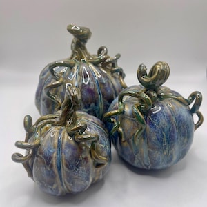 May include: Three ceramic pumpkins in shades of blue, purple, and gold. Each pumpkin has a textured surface and a twisted stem with tendrils. The pumpkins are arranged in a group, with varying sizes.
