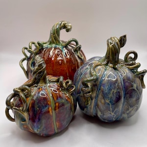 May include: Three ceramic pumpkins in different sizes and colours. The pumpkins have a glossy finish with shades of red, purple, and blue. Each pumpkin features a detailed, twisted stem and vine-like accents, ideal for autumn decor.