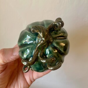 May include: A green ceramic pumpkin with a brown stem and vine detail. The pumpkin has a glossy finish and is a decorative item.