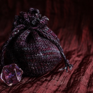 May include: A purple and grey knitted drawstring bag with a clear iridescent dice with purple numbers inside.
