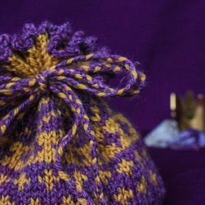 May include: A knitted drawstring bag in shades of purple and gold. The bag features a diamond pattern and a matching drawstring closure. The background is a solid purple, with a blurred gold crown and a blue crystal.
