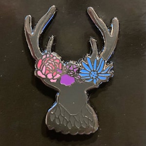 May include: Black enamel pin of a deer head with antlers, wearing a flower crown with pink, purple, and blue flowers.