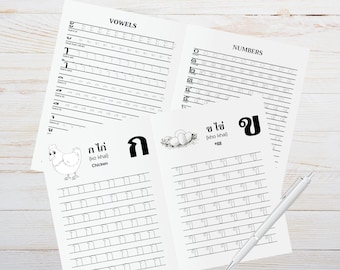 May include: White paper learning sheets with Thai script, including vowels, numbers, and words like "chicken" and "egg." A white pen rests on the wooden surface. The sheets feature lines for practicing writing.