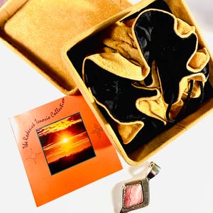 May include: An open, square, tan leather box with a black interior and a pair of tan gloves. A small orange card with a sunset photo and text "The Roderick Tenorio Collection" is next to a silver pendant with a pink stone.