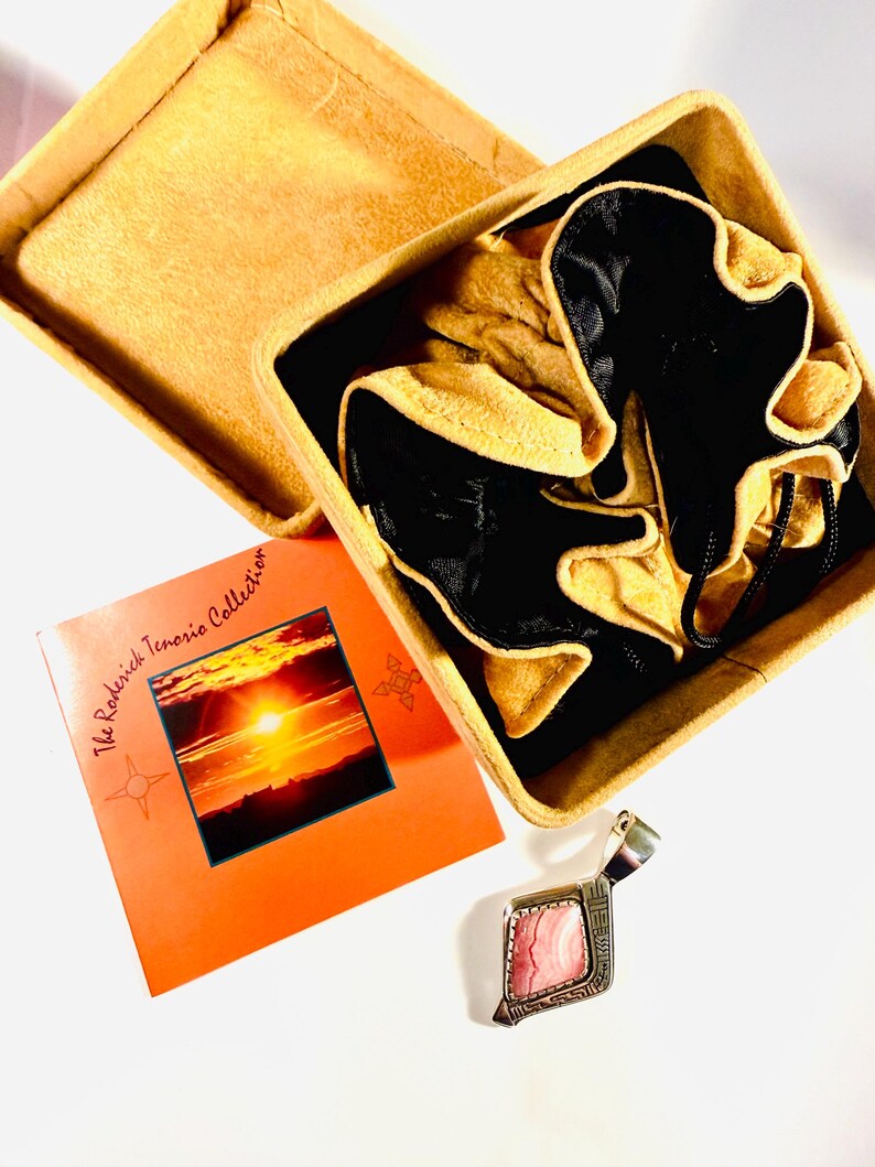 May include: An open, square, tan leather box with a black interior and a pair of tan gloves. A small orange card with a sunset photo and text "The Roderick Tenorio Collection" is next to a silver pendant with a pink stone.