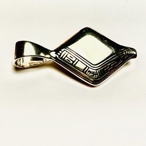 May include: A silver pendant with a geometric design. The pendant has a rectangular shape with a loop for a chain. The surface is decorated with engraved patterns and lines.